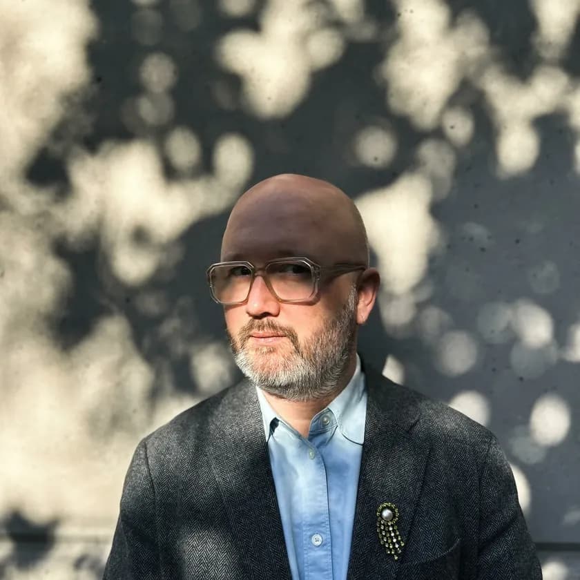 A man with glasses and a beard sits against a dappled light background, wearing a blue shirt and dark blazer with a decorative pin.