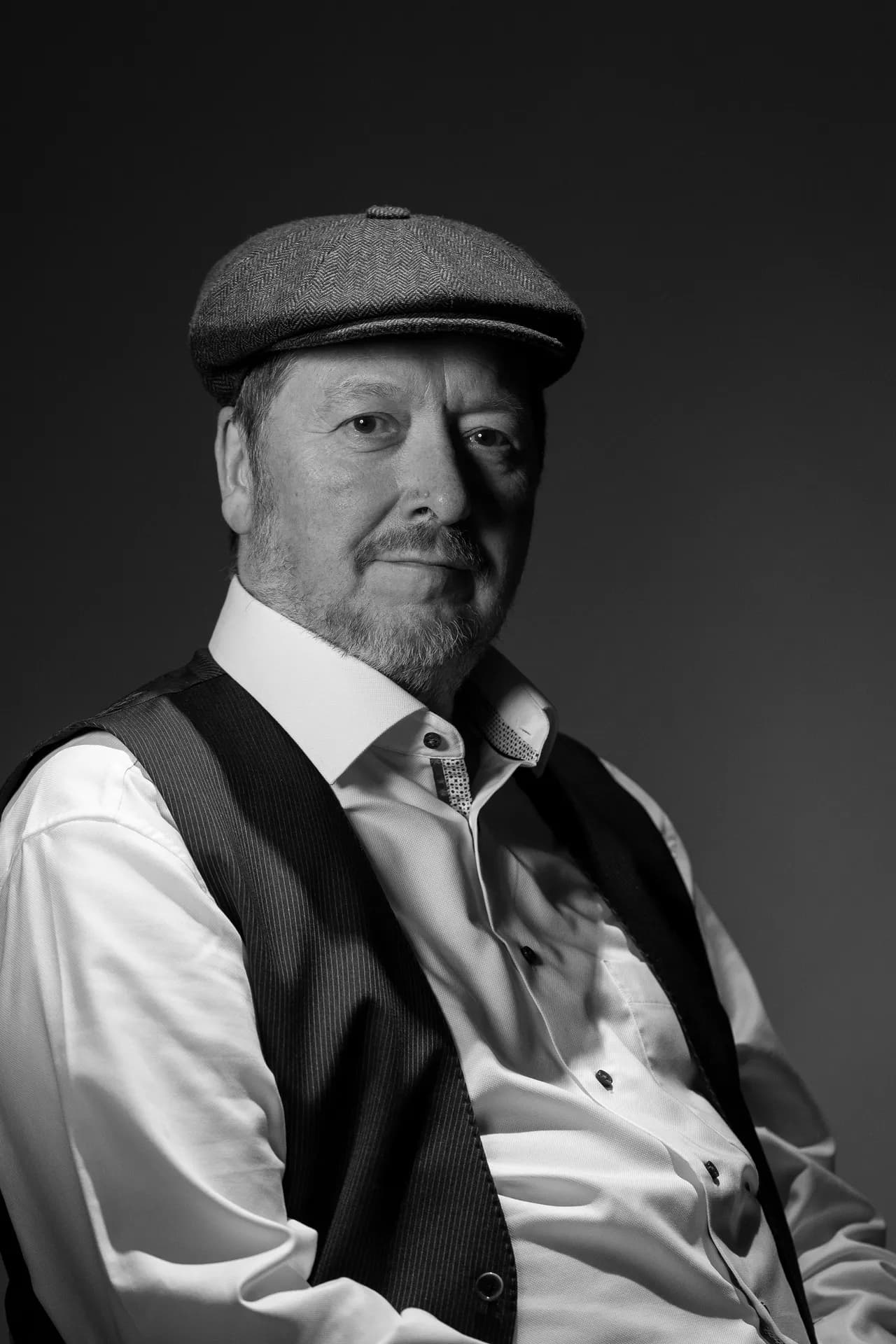 A man wearing a flat cap and a vest over a white shirt, posed in a seated position against a dark background in black and white.