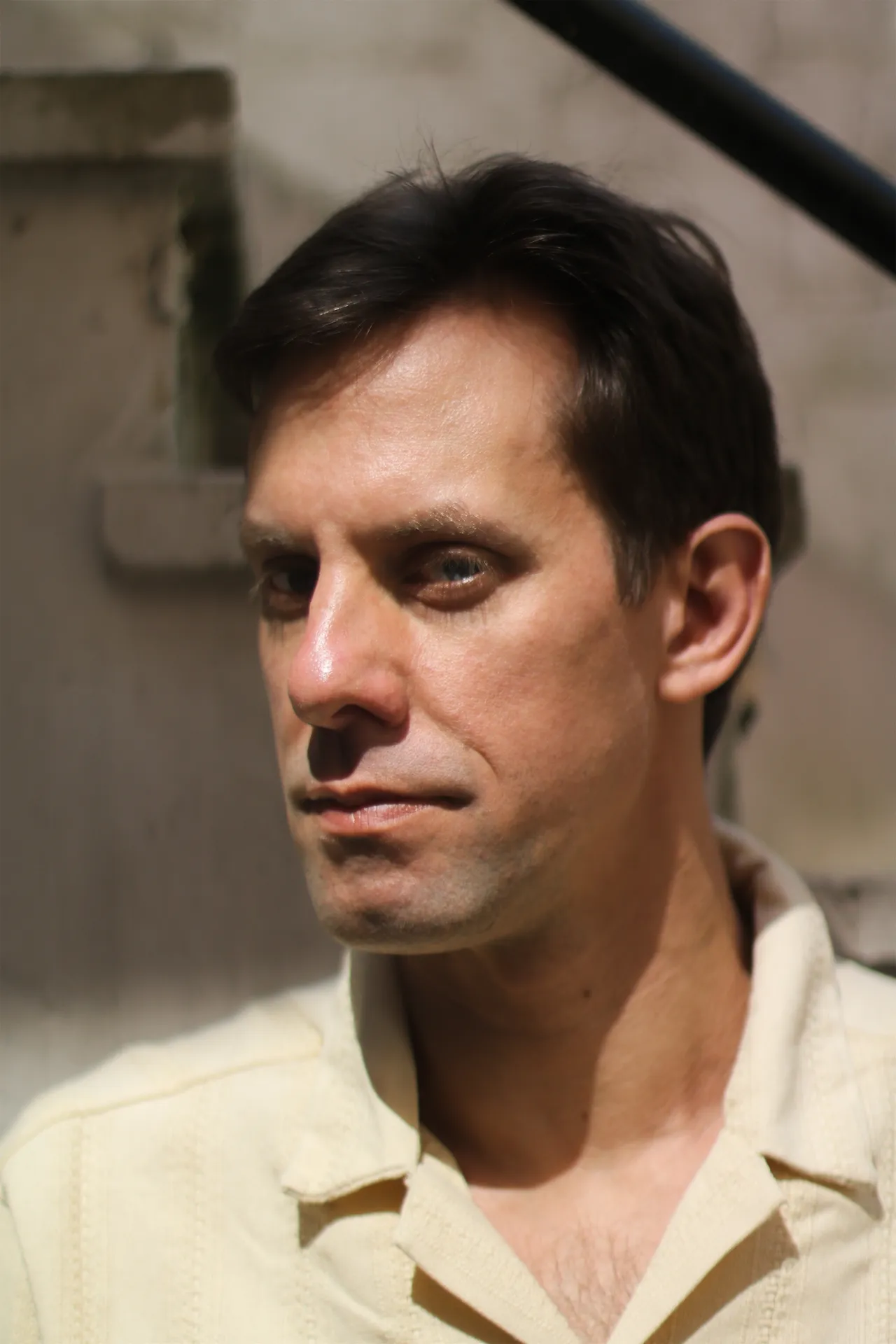 A man in a yellow shirt is sitting outdoors, lit by sunlight, looking to the side with a thoughtful expression.