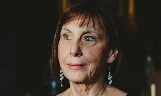 A woman in a blue top, wearing earrings and a necklace, looks to the side in soft lighting.