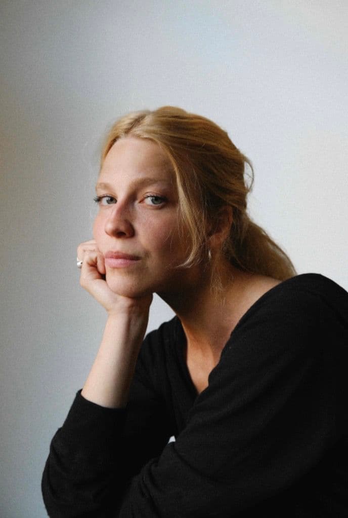 A person with long blonde hair and a black top rests their chin on their hand, gazing thoughtfully at the camera against a plain background.