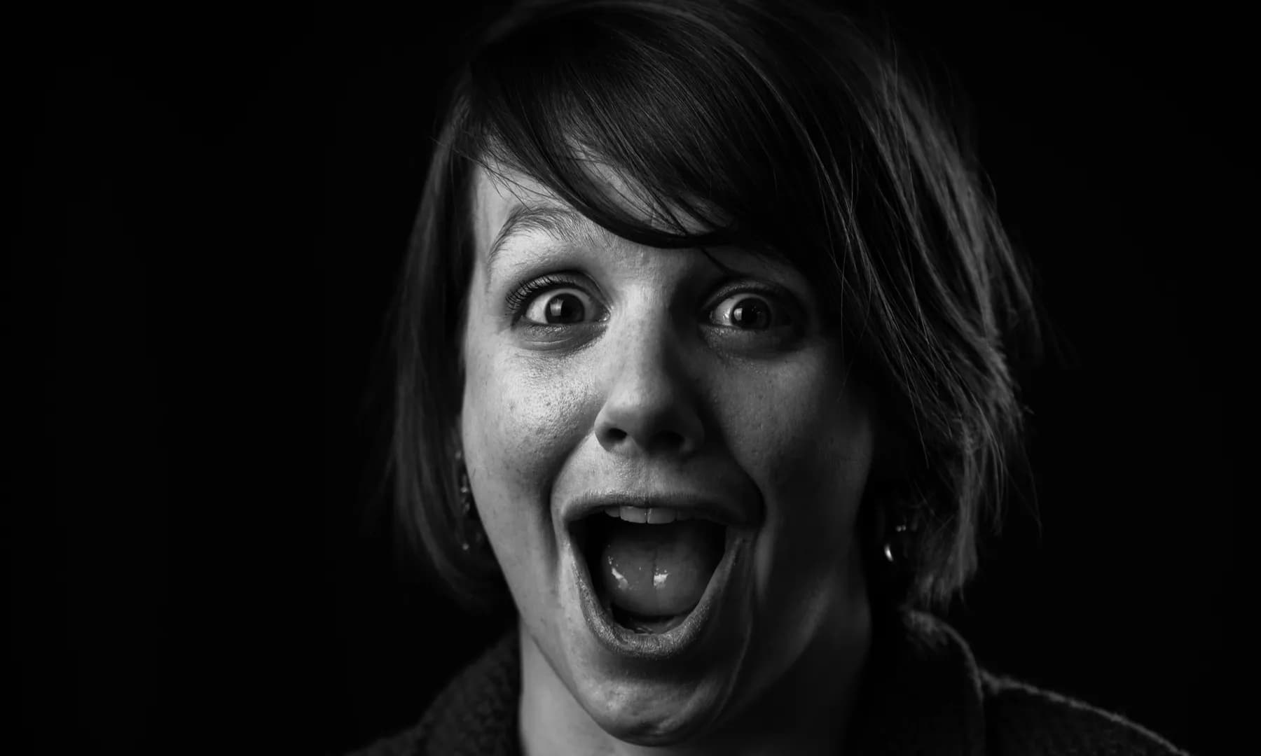Black and white portrait of a surprised person with an open mouth and wide eyes, set against a dark background.