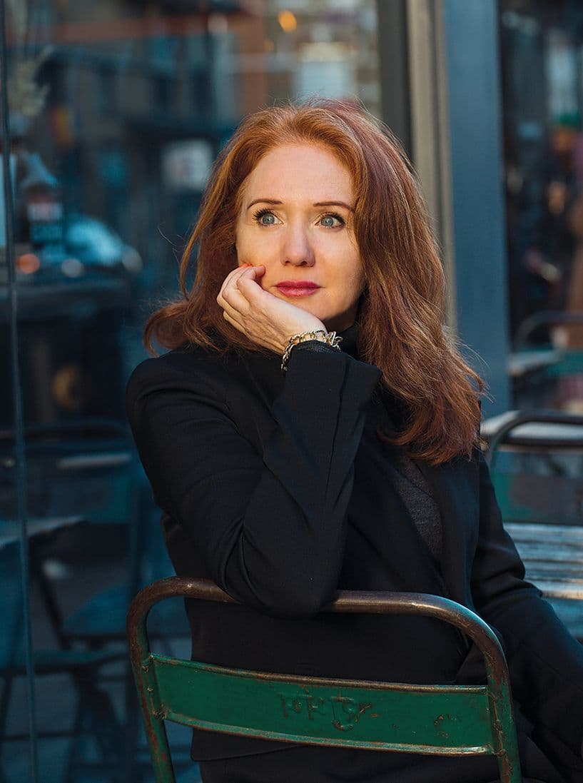 Woman with red hair in a black blazer sits pensively on a green chair outdoors, resting her chin on her hand.
